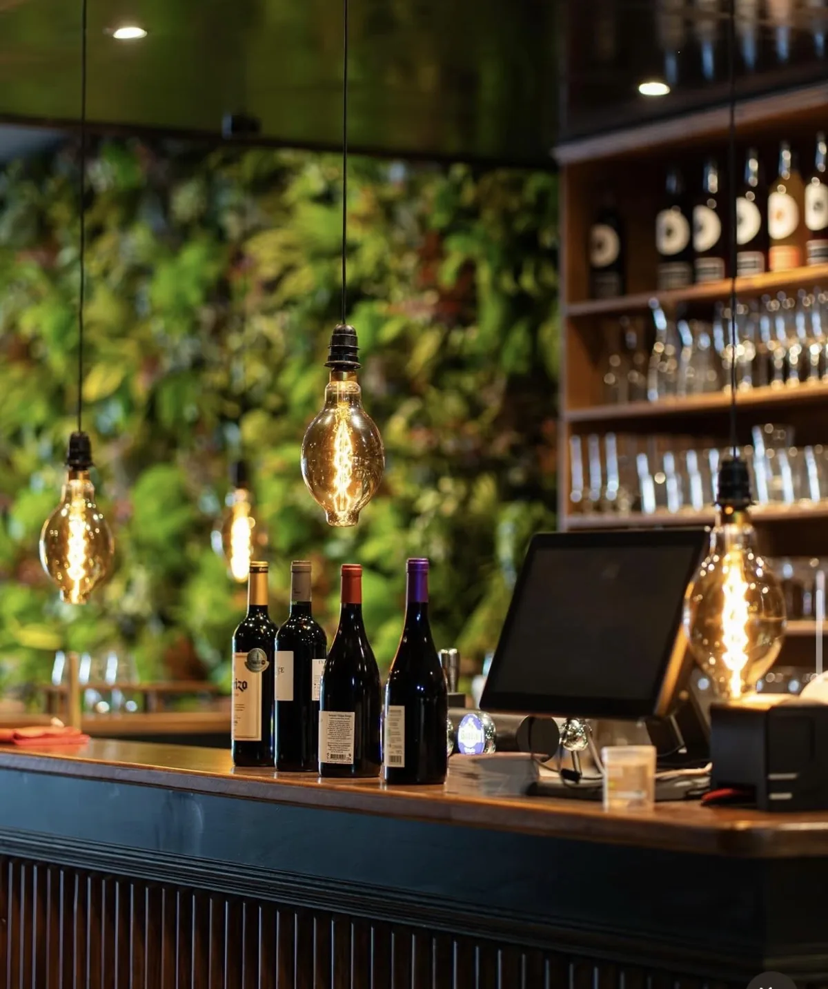 Comptoir du Manhattan Grill : bouteilles de vin, ampoules à filament Edison, mur végétal et étagère à verres.
